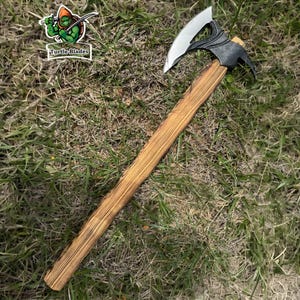 May include: A tomahawk axe with a wooden handle and a black and silver head. The axe is lying on a bed of grass. The text "Turtle Blades" is visible on a white rectangle.