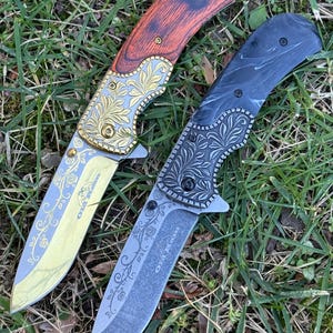 Op de afbeelding: Twee inklapbare messen met florale gravures op de bladen en kropstukken. Het linker mes heeft een roodbruin houten handvat en een goudkleurig blad. Het rechter mes heeft een zwart gemarmerd handvat en een donkergrijs blad. Tekst OLA R. zichtbaar.