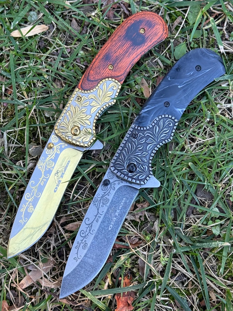 Op de afbeelding: Twee inklapbare messen met florale gravures op de bladen en kropstukken. Het linker mes heeft een roodbruin houten handvat en een goudkleurig blad. Het rechter mes heeft een zwart gemarmerd handvat en een donkergrijs blad. Tekst OLA R. zichtbaar.
