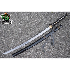 May include: A katana sword with a silver blade, a black handle wrapped in white cord, and a black scabbard featuring a grey cord and gold-coloured emblem. The blade has a visible engraving near the hilt. A logo with a turtle and the text Turtle Blades.