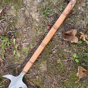 Pu&ograve; includere: Un'arma inastata con una lama in acciaio lucido e un'asta in legno. La lama ha una forma curva distintiva con due punte rivolte verso l'esterno. L'asta &egrave; marrone chiaro con bande scure decorative e piccoli borchie metalliche.