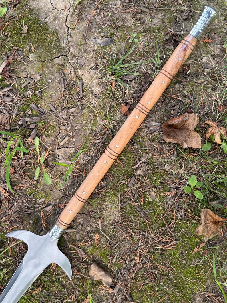 Pu&ograve; includere: Un'arma inastata con una lama in acciaio lucido e un'asta in legno. La lama ha una forma curva distintiva con due punte rivolte verso l'esterno. L'asta &egrave; marrone chiaro con bande scure decorative e piccoli borchie metalliche.