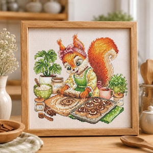 May include: A framed cross-stitch embroidery featuring an orange squirrel wearing a green apron and a pink headband, rolling dough on a wooden board. The scene includes a baking tray of cinnamon rolls, a flour container, and potted plants.