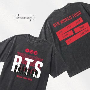 Peut inclure: T-shirts gris foncé avec un motif graphique. Le devant du t-shirt présente le mot "BTS" en blanc, avec un motif de pagode rouge et le texte "WORLD TOUR 2026". Le dos du t-shirt affiche "BTS WORLD TOUR" et les noms des membres.