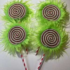 May include: Four green fuzzy lollipop decorations with red and white candy cane swirl designs. The lollipops are attached to red and white striped sticks.