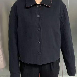 May include: A black jacket with a classic collar and button-down front. The collar has a red and white patterned trim. The jacket is worn with black pants. The jacket has long sleeves and a cropped length.
