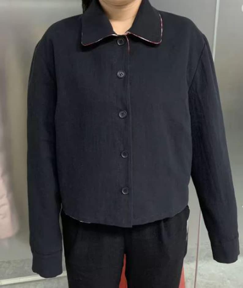 May include: A black jacket with a classic collar and button-down front. The collar has a red and white patterned trim. The jacket is worn with black pants. The jacket has long sleeves and a cropped length.