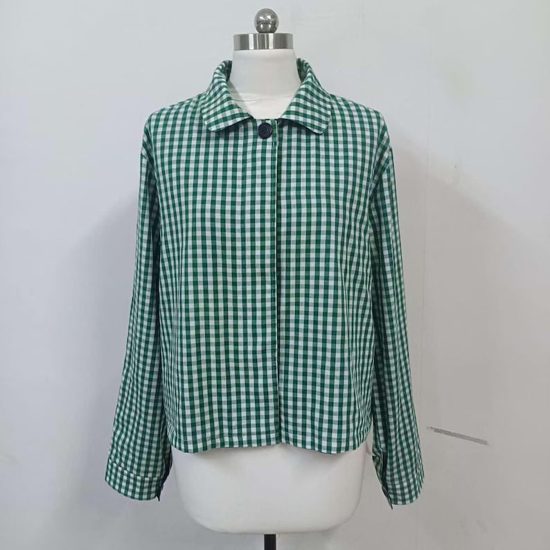 May include: A green and white gingham-patterned jacket with long sleeves and a collar. The jacket has a single black button closure and is displayed on a mannequin. The fabric has a classic checkered design, offering a casual yet stylish look.