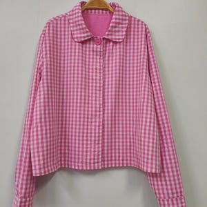 May include: A pink and white gingham button-down shirt with long sleeves and a rounded collar. The shirt is hanging on a wooden hanger against a plain white background. The shirt has a single pink button.