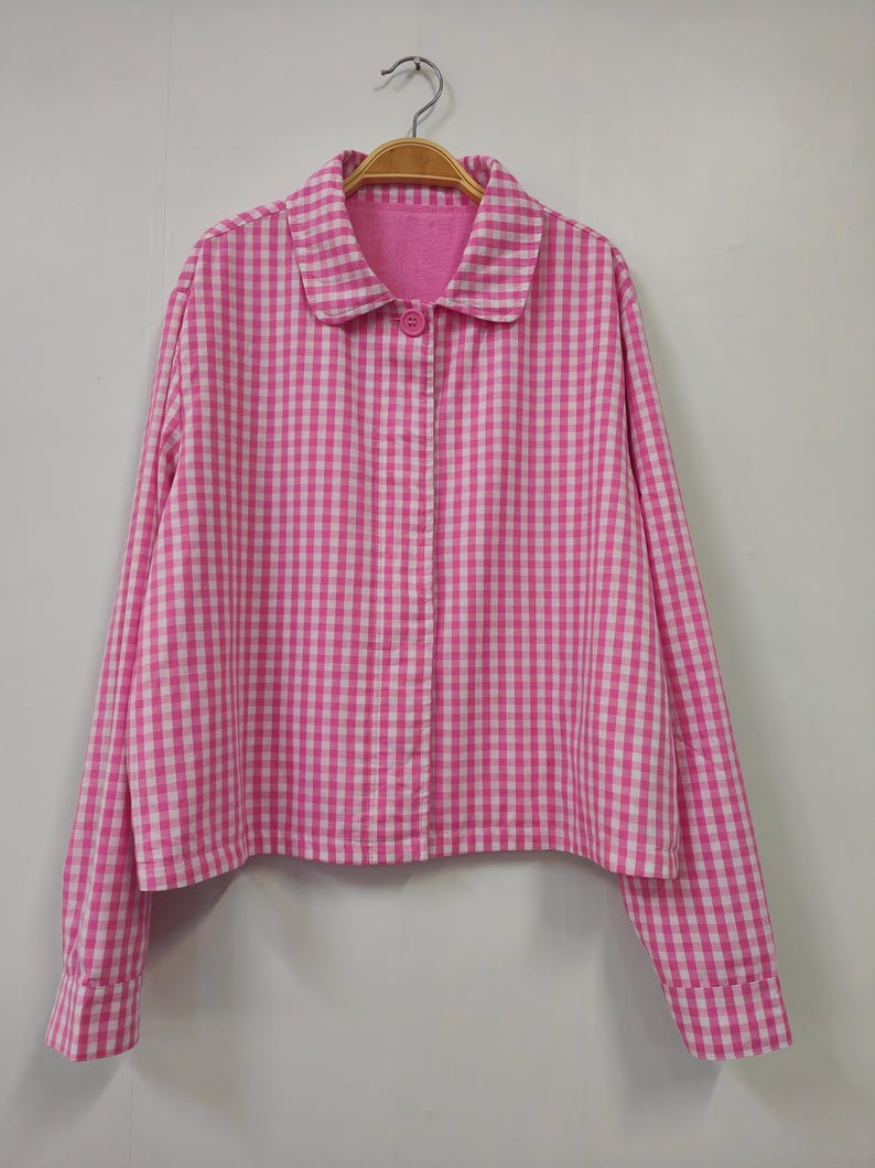 May include: A pink and white gingham button-down shirt with long sleeves and a rounded collar. The shirt is hanging on a wooden hanger against a plain white background. The shirt has a single pink button.