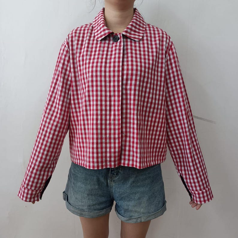 May include: A red and white gingham-patterned jacket with long sleeves and a button closure. The jacket has a classic collar and is paired with denim shorts. The overall style is casual and preppy.
