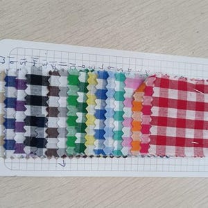 May include: A collection of fabric swatches in various colors and patterns, including gingham and plaid. The swatches are arranged on a white grid paper, showcasing a range of hues from purple and black to red and white. The edges of the fabric are jagged.