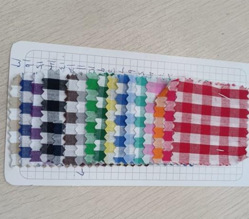 May include: A collection of fabric swatches in various colors and patterns, including gingham and plaid. The swatches are arranged on a white grid paper, showcasing a range of hues from purple and black to red and white. The edges of the fabric are jagged.