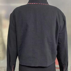 May include: Black jacket with a collar and long sleeves. The collar has a thin red and white stripe. The sleeves have a red and white stripe detail at the cuffs. The jacket is cropped at the waist.