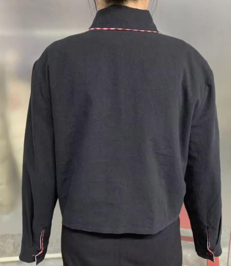May include: Black jacket with a collar and long sleeves. The collar has a thin red and white stripe. The sleeves have a red and white stripe detail at the cuffs. The jacket is cropped at the waist.