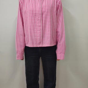 May include: A pink and white checkered jacket with a button closure and a rounded collar. The jacket is paired with dark blue jeans. The long-sleeved jacket has a classic gingham pattern.