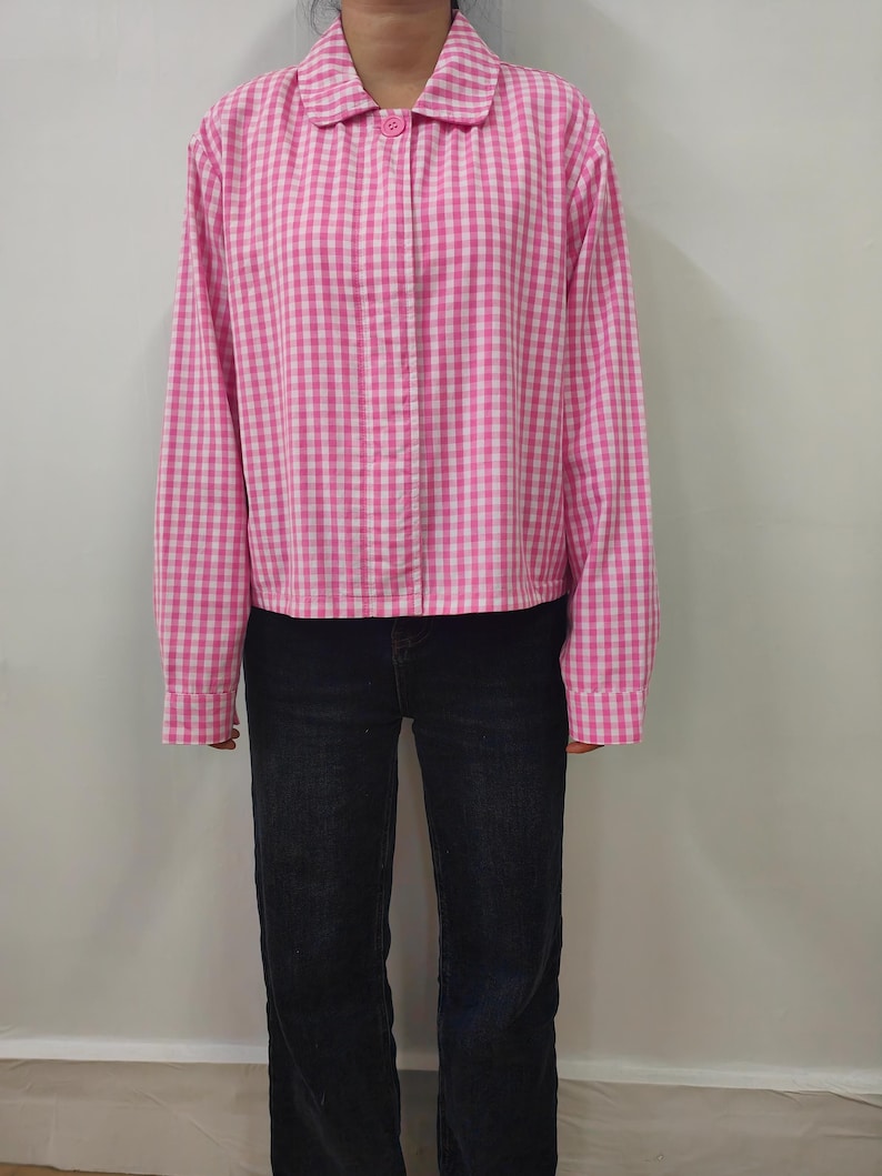May include: A pink and white checkered jacket with a button closure and a rounded collar. The jacket is paired with dark blue jeans. The long-sleeved jacket has a classic gingham pattern.