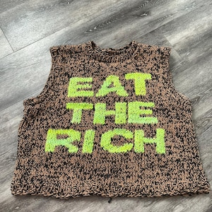 Könnte beinhalten: Ein brauner und schwarzer Strickpullover ohne Ärmel mit der Aufschrift "EAT THE RICH" in grüner Schrift.