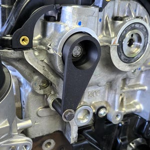 May include: Close-up of a black metal engine bracket with a bolt and nut. The bracket is attached to a silver engine block with a black plastic cover. The engine block has a black plastic cover with a gold screw. The engine block has a silver metal pulley with a black rubber belt.