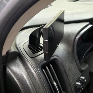May include: A black car phone mount attached to the air vent in a car. The mount is adjustable and can be rotated to different angles.