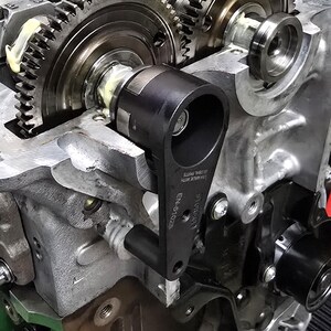 Pu&ograve; includere: Primo piano di un blocco motore argentato con un tenditore della catena di distribuzione nero e una guida della catena di distribuzione nera. Il blocco motore ha un ingranaggio argentato e un ingranaggio nero. Il tenditore della catena di distribuzione ha la scritta "EN-5102S" stampata su di esso.