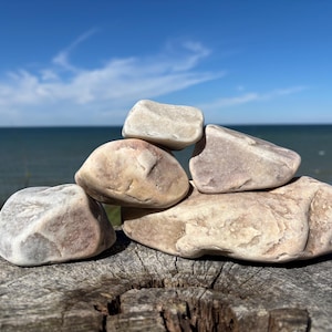 May include: A stack of five smooth, rounded river stones in shades of beige, tan, and light gray. The stones are arranged in a balanced formation, with one large base stone supporting four smaller, irregularly shaped rocks on top.
