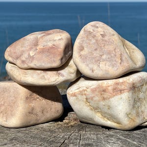 May include: A stack of four smooth, rounded stones with a pale beige and light pinkish hue. The stones are arranged in a balanced formation, featuring natural textures, subtle mineral veins, and irregular shapes with soft, weathered edges.