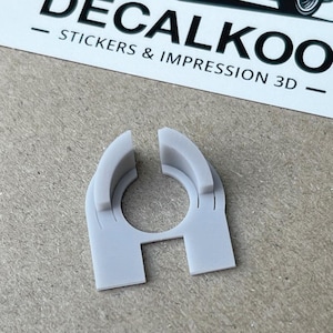 Puede incluir: Una pieza de plástico gris impresa en 3D con una abertura circular y dos ranuras verticales. La pieza está diseñada para ajustarse alrededor de un objeto cilíndrico.