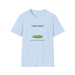 Puede incluir: Una camiseta de manga corta de color azul claro con un gráfico de un hoyo de golf verde con una bandera blanca. Sobre el gráfico aparece el texto "I HATE GOLF!" y debajo del gráfico el texto "Wanna Play Tomorrow?" en una fuente negra sencilla.