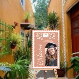 May include: A welcome sign for a graduation party with a peach background and white decorative border. The sign features a photo of a graduate wearing a cap and gown and the text "Isabella's Graduation Party" and "Welcome".