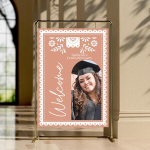 May include: A peach-colored welcome sign with a white border and a white cutout design. The sign says "Welcome" and "Isabella's Graduation Party". A photo of a graduate in a cap and gown is in the center of the sign.