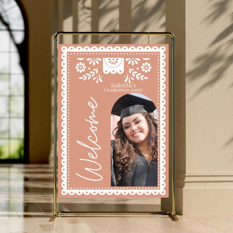 May include: A peach-colored welcome sign with a white border and a white cutout design. The sign says "Welcome" and "Isabella's Graduation Party". A photo of a graduate in a cap and gown is in the center of the sign.