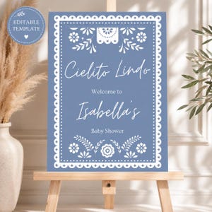 May include: A blue baby shower sign with white floral and decorative border. The sign reads "Cielito Lindo, Welcome to Isabella's, Baby Shower". The sign is on a wooden easel.