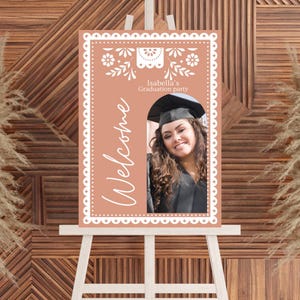 May include: A peach-colored welcome sign with a white border and a white cutout design. The sign says "Welcome" and "Isabella's Graduation Party". A photo of a graduate in a cap and gown is in the center of the sign.
