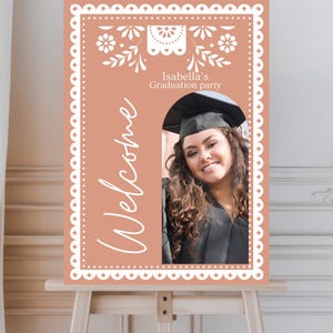 May include: A peach-colored welcome sign with a white scalloped border and floral design. The sign says "Welcome" and "Isabella's Graduation Party". There is a cutout for a photo of a graduate wearing a cap and gown.