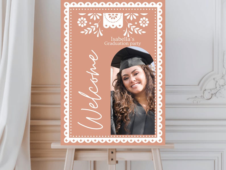 May include: A peach-colored welcome sign with a white scalloped border and floral design. The sign says "Welcome" and "Isabella's Graduation Party". There is a cutout for a photo of a graduate wearing a cap and gown.