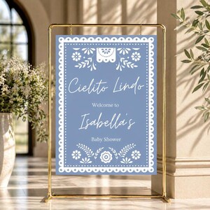 May include: A baby shower sign with a gold metal frame. The sign has a blue background with white floral and decorative elements. The text reads "Cielito Lindo", "Welcome to Isabella's", and "Baby Shower".