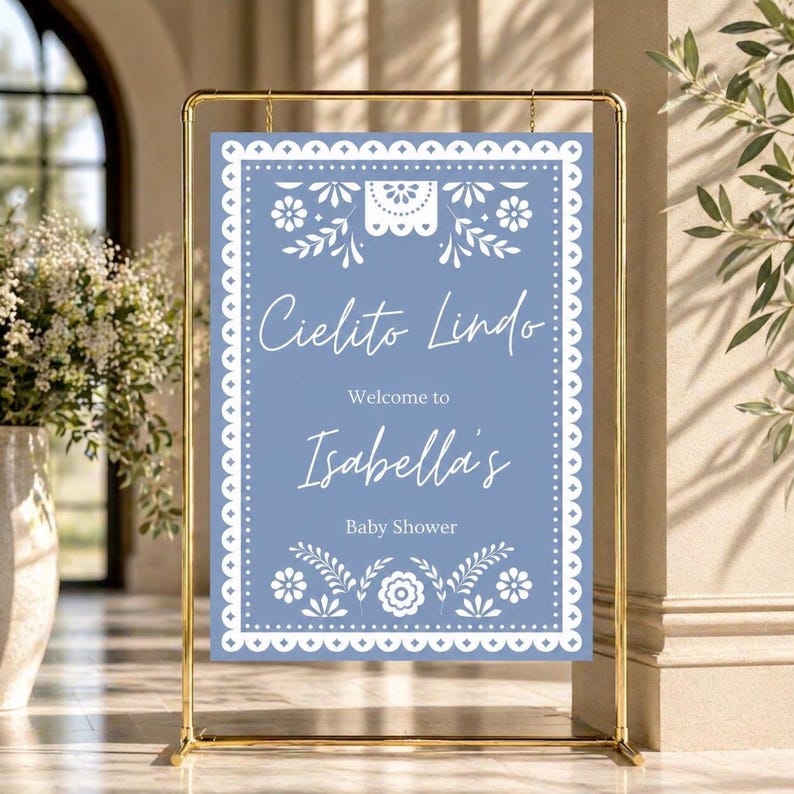 May include: A baby shower sign with a gold metal frame. The sign has a blue background with white floral and decorative elements. The text reads "Cielito Lindo", "Welcome to Isabella's", and "Baby Shower".