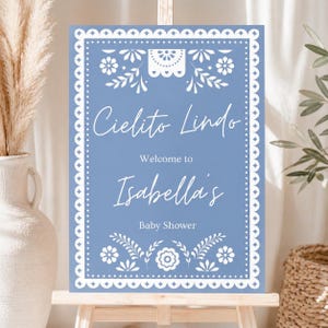May include: A baby shower welcome sign with a light blue background and white decorative border. The sign reads "Cielito Lindo" and "Welcome to Isabella's Baby Shower" in elegant script. The design includes floral and decorative elements.