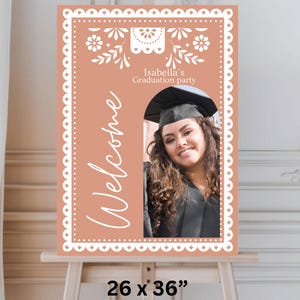 May include: A peach-colored welcome sign with a white decorative border and a cutout for a photo. The sign says "Isabella's Graduation Party" and "Welcome" in white script. The sign is 26 inches by 36 inches.