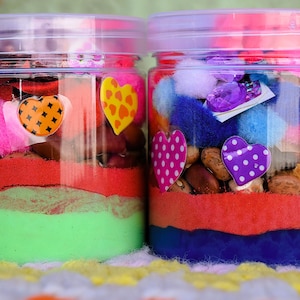 May include: Two clear plastic jars filled with layers of colorful sand, beans, and decorative elements. Each jar has a clear lid and is adorned with heart-shaped stickers and fluffy embellishments in various colors. The jars are on a colorful, patterned surface.