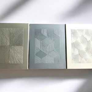 Pode incluir: Três peças de arte de parede retangulares com diferentes padrões geométricos em relevo. A peça da esquerda é bege com um padrão radiante. A peça do centro é azul claro com um padrão geométrico de quadrados e linhas. A peça da direita é branca com um padrão de círculos sobrepostos.