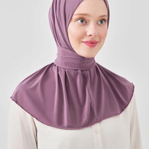 May include: A mauve-coloured hijab with a fitted head covering and a pleated neck-covering piece that drapes over the shoulders. The fabric is smooth and opaque, providing full coverage for the head and neck area in a solid, muted purple tone.