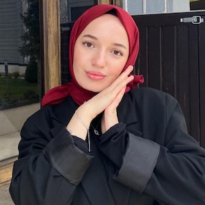 Puede incluir: Una mujer con un pañuelo de cabeza burdeos y una chaqueta negra con un cinturón negro y una hebilla dorada. Está posando con las manos juntas delante de ella.