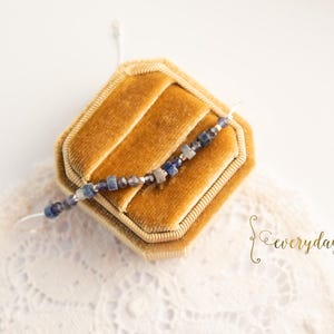 Op de afbeelding: Een delicate armband met blauwe en zilveren kralen ligt op een gouden fluwelen sieradendoosje. De doos staat op een wit kanten kleedje. Het woord "everyday" is cursief geschreven.