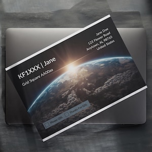 May include: A black and white business card featuring a graphic of Earth with a sunrise. The card includes the name "Jane Doe", address, and the text "KF1XXX | Jane" and "Grid Square AA00aa". The card is on a laptop.