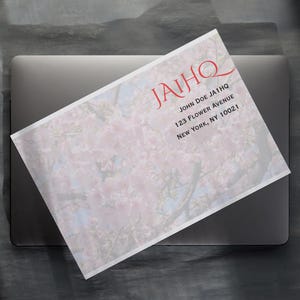 May include: A white envelope with a floral design and the text "JAIHQ" in red, along with an address. The envelope is placed on top of a closed laptop, with a dark gray background.