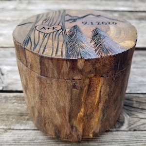 K&ouml;nnte beinhalten: Eine Holzkiste f&uuml;r Ringe mit einem Deckel, der mit einem Herzen, den Initialen "A+K", zwei Kiefern, einer Bergkette und dem Datum "9.1.2019" graviert ist.