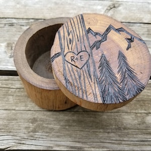 K&ouml;nnte beinhalten: Holzschachtel f&uuml;r Ringe mit einem Herz und den Initialen R+E, die in den Deckel graviert sind. Der Deckel zeigt au&szlig;erdem eine Bergkette und zwei Kiefern.
