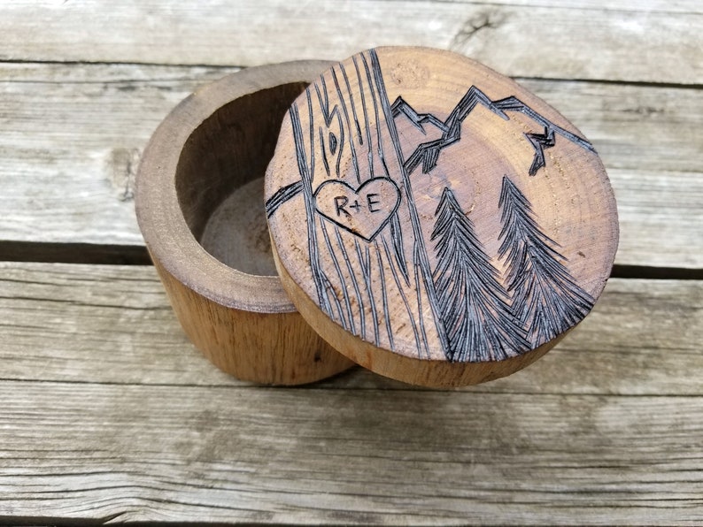 K&ouml;nnte beinhalten: Holzschachtel f&uuml;r Ringe mit einem Herz und den Initialen R+E, die in den Deckel graviert sind. Der Deckel zeigt au&szlig;erdem eine Bergkette und zwei Kiefern.